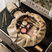 Borsa trucco beige aperta su tavolo nero, contenente cosmetici vari: rossetti, pennelli, ombretti. Sfondo con poster artistico. Accessori bellezza.