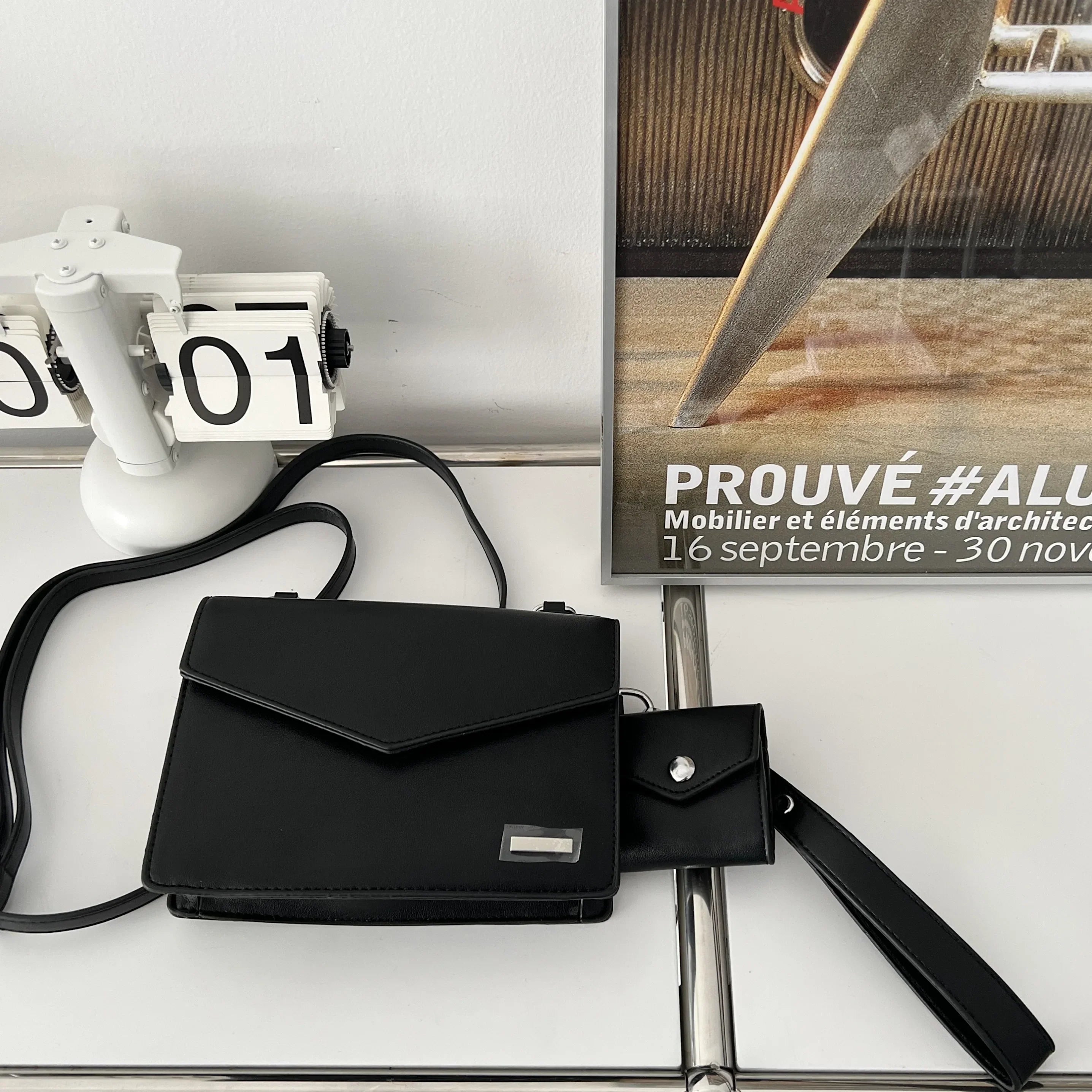 Borsa a tracolla nera elegante su tavolo bianco, accanto a calendario e poster di design. Accessorio moda donna, stile minimalista e moderno.