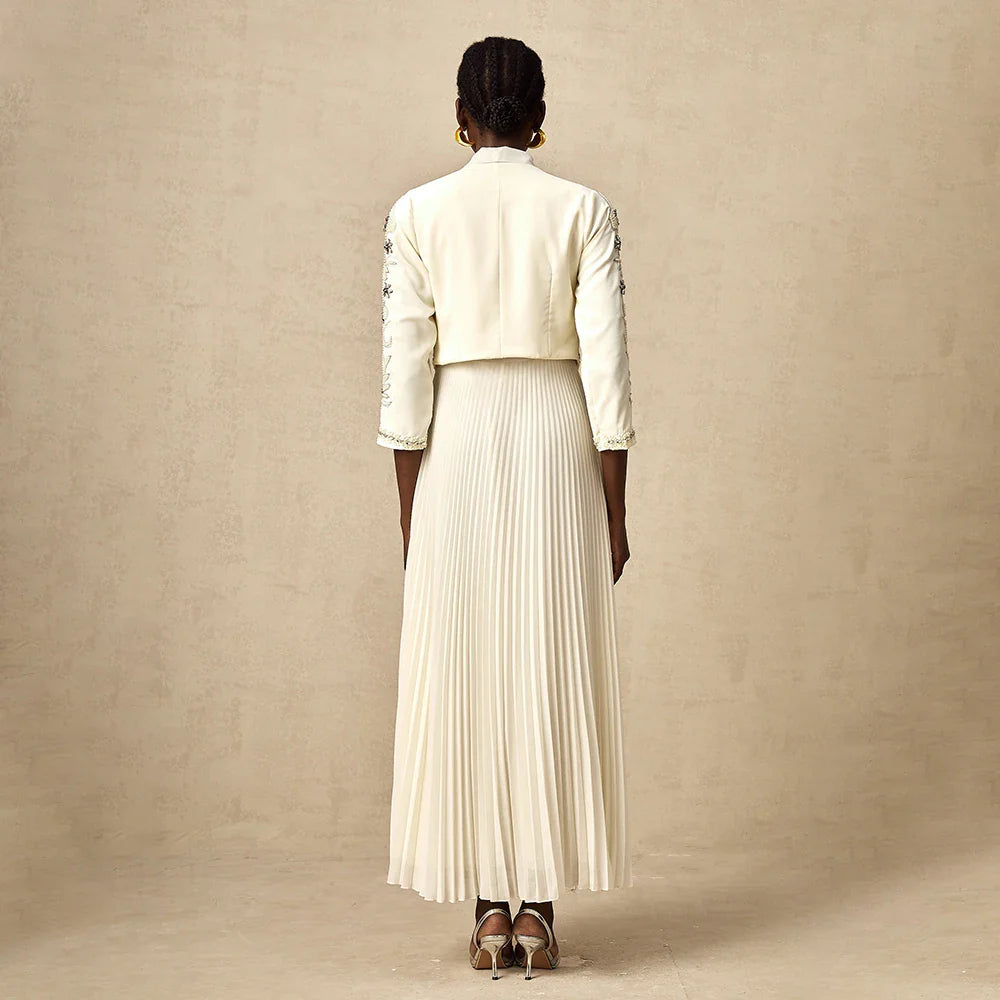 Donna di spalle con abito lungo plissettato bianco, giacca corta ricamata, orecchini dorati, su sfondo neutro. Moda elegante, abbigliamento femminile.