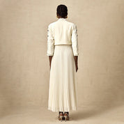 Donna di spalle con abito lungo plissettato bianco, giacca corta ricamata, orecchini dorati, su sfondo neutro. Moda elegante, abbigliamento femminile.