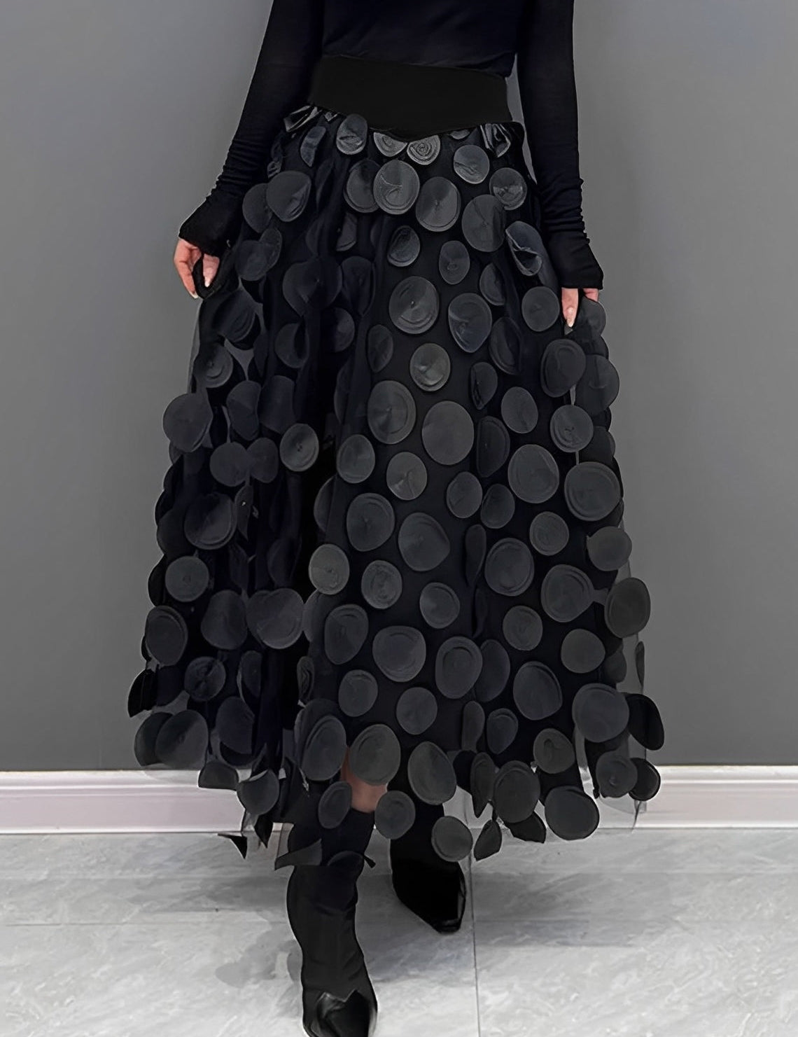 Gonna lunga nera con cerchi decorativi, indossata con top nero a maniche lunghe e stivali a punta. Moda elegante, abbigliamento femminile chic.