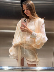Donna in ascensore indossa abito elegante color crema, maniche ampie, tessuto leggero. Scatta selfie con smartphone. Moda femminile, abbigliamento chic.