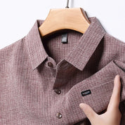 Camicia uomo a maniche lunghe, tessuto a quadri marrone, colletto classico, bottoni frontali, etichetta "Fashion", appesa su gruccia di legno.