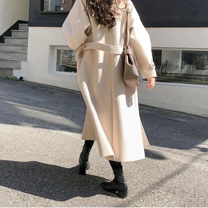 Donna con cappotto beige lungo, borsa a tracolla marrone e stivali neri, cammina su strada urbana. Moda autunnale elegante e casual.