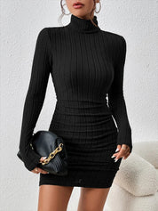 Abito nero a coste, maniche lunghe, collo alto, aderente, elegante. Donna con borsa a catena dorata. Moda autunno inverno.