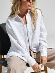 Donna con camicia bianca di cotone, seduta su sedia nera, indossa occhiali da sole. Moda casual elegante, abbigliamento donna, stile estivo.