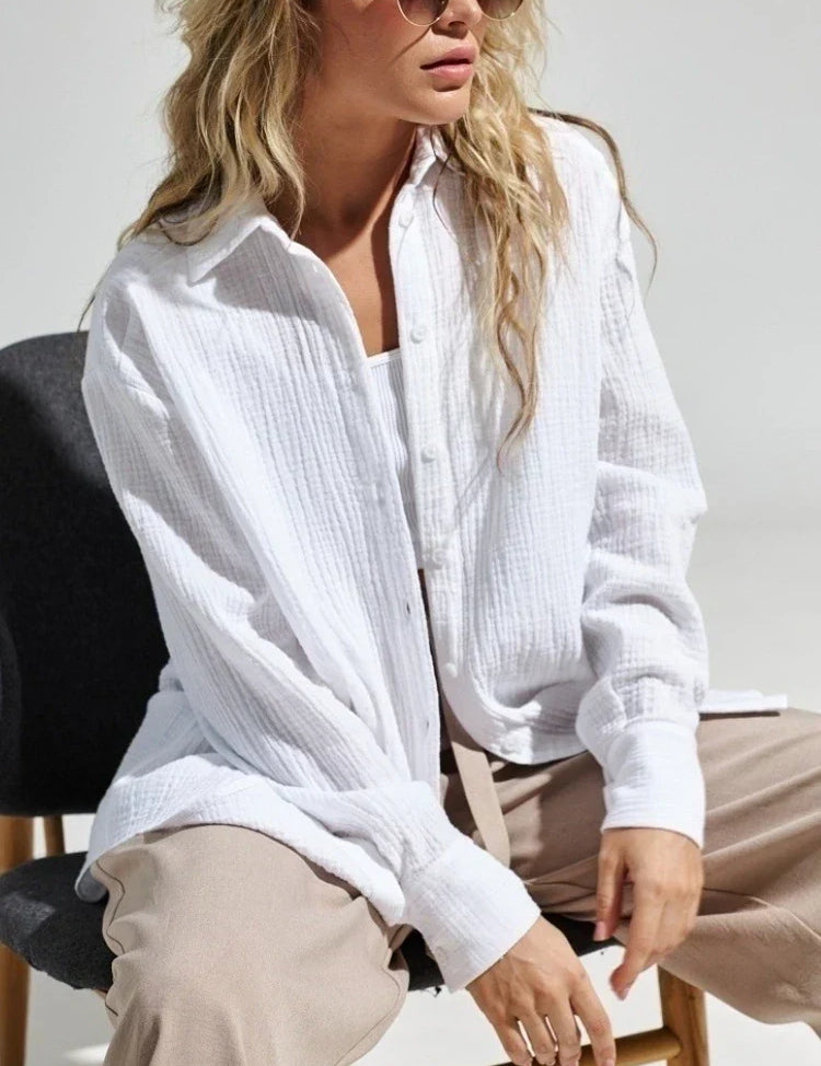 Donna con camicia bianca di cotone, seduta su sedia nera, indossa occhiali da sole. Moda casual elegante, abbigliamento donna, stile estivo.