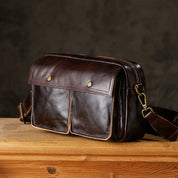 Borsa a tracolla in pelle marrone scuro su tavolo di legno, con doppie tasche frontali e chiusura a zip. Accessorio moda elegante e funzionale.
