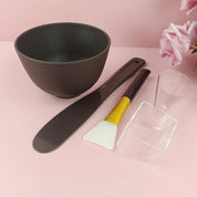 Set ciotola e accessori per maschera viso su sfondo rosa, include spatola, pennello, misurino e ciotola nera. Ideale per skincare e bellezza.