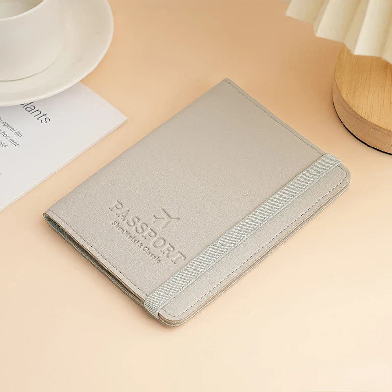 Custodia passaporto in pelle grigia su tavolo beige, con elastico e tazza bianca. Accessorio da viaggio elegante e funzionale.