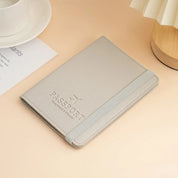 Custodia porta passaporto in pelle grigia su tavolo beige, con elastico e scritta "Passport". Accessorio da viaggio elegante e funzionale.