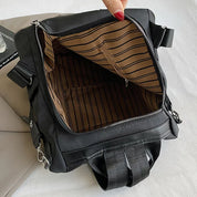 Zaino nero aperto con interno a righe marroni, tasca con zip, manico superiore e spallacci regolabili. Design moderno e funzionale per viaggi.