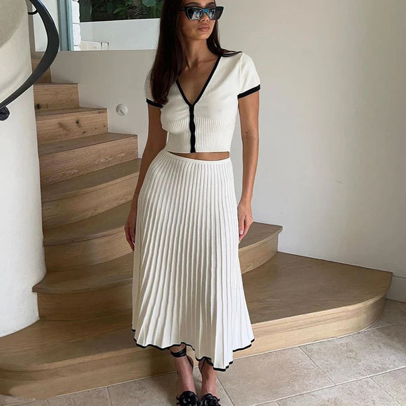 Donna con abito bianco a coste, gonna plissettata, top corto, occhiali da sole neri, su scala in legno. Moda estiva elegante, stile casual chic.