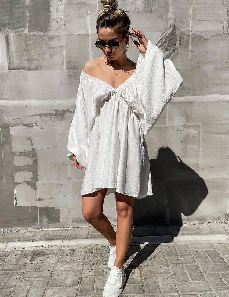 Donna con abito bianco estivo, spalle scoperte, maniche ampie, occhiali da sole, scarpe da ginnastica bianche, posa su sfondo urbano grigio.