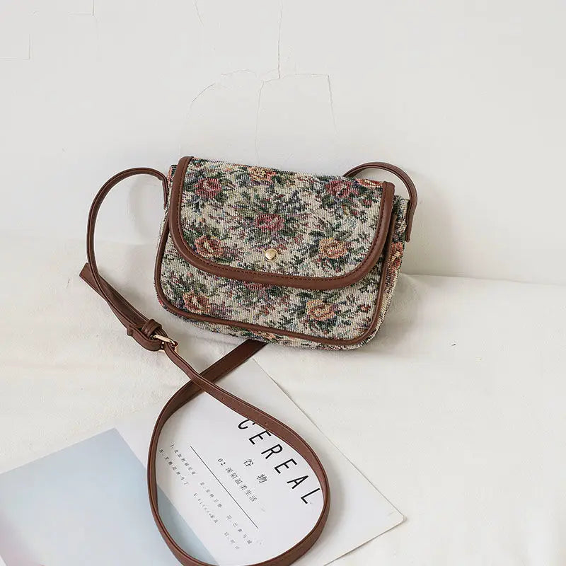 Borsa a tracolla vintage floreale in tessuto jacquard, dettagli in pelle marrone, chiusura a bottone, su sfondo bianco. Accessorio moda elegante.