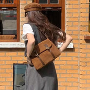 Donna con borsa a tracolla marrone in pelle scamosciata, abbigliamento autunnale, cappello coordinato, sfondo di mattoni. Moda casual femminile.