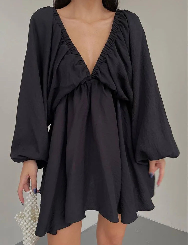 Abito nero corto con scollo a V profondo e maniche lunghe a sbuffo, tenuto da una donna. Elegante e moderno, perfetto per serate e occasioni speciali.