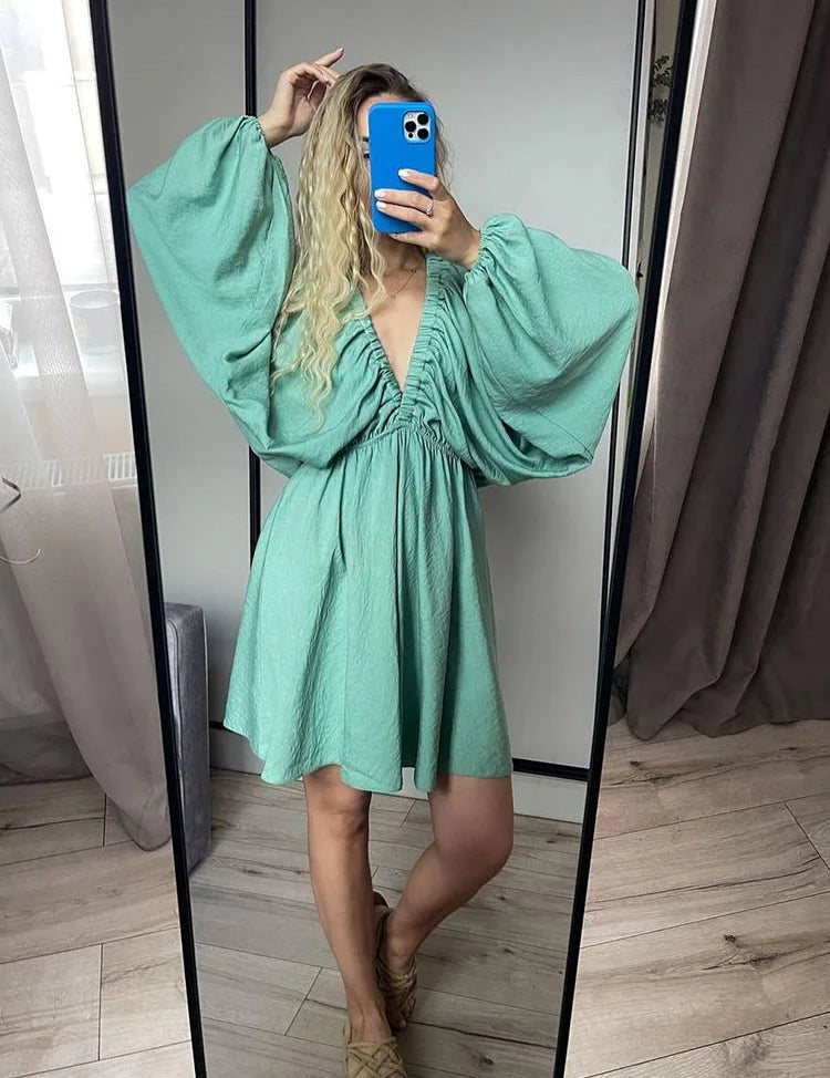 Donna in abito verde con maniche a sbuffo, scollo a V, posa davanti a uno specchio. Moda femminile, abbigliamento estivo elegante.