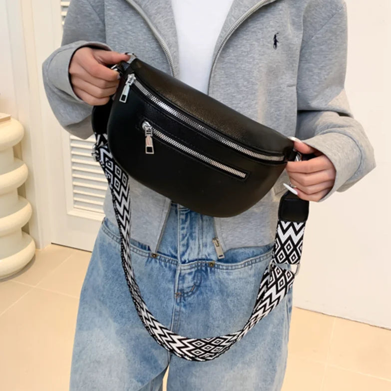 Borsa a tracolla nera in pelle con cerniere argentate, indossata su giacca grigia e jeans. Cinturino regolabile con motivo geometrico bianco e nero.