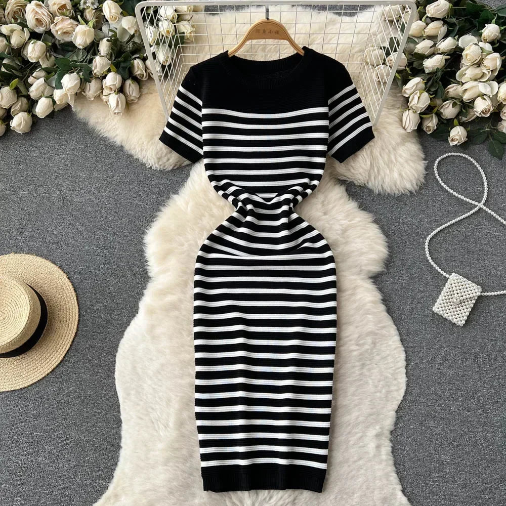 Abito a righe bianche e nere su tappeto bianco, circondato da rose bianche e cappello di paglia. Elegante vestito estivo, moda donna.