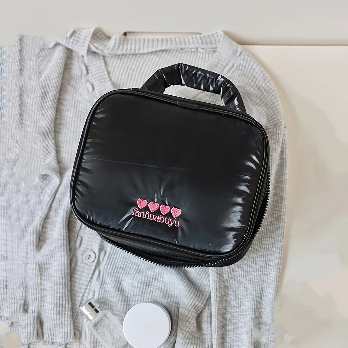 Borsa cosmetica nera su maglione grigio, con logo ricamato rosa. Ideale per viaggi, organizzazione trucco, accessori bellezza.
