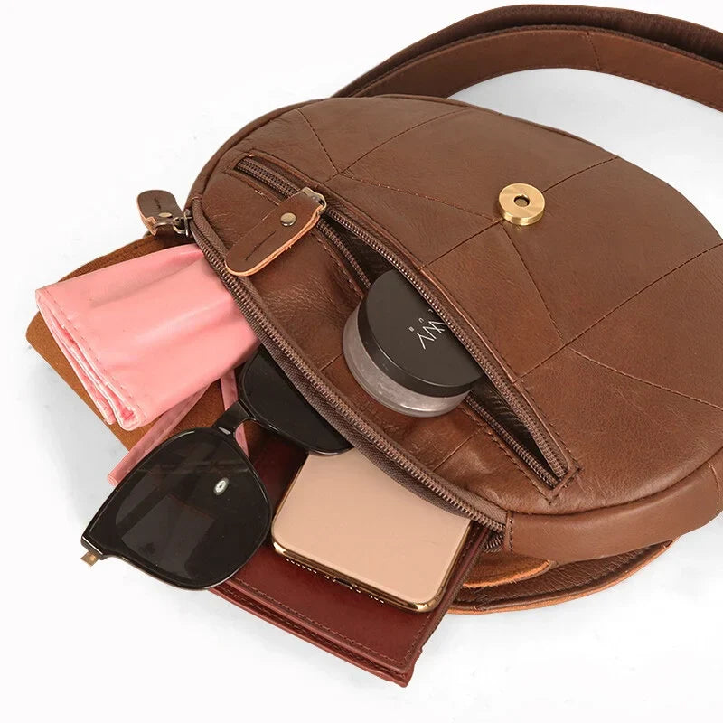 Borsa a tracolla in pelle marrone con cerniera aperta, contenente occhiali da sole, smartphone, rossetto e fazzoletto rosa. Accessori moda donna.