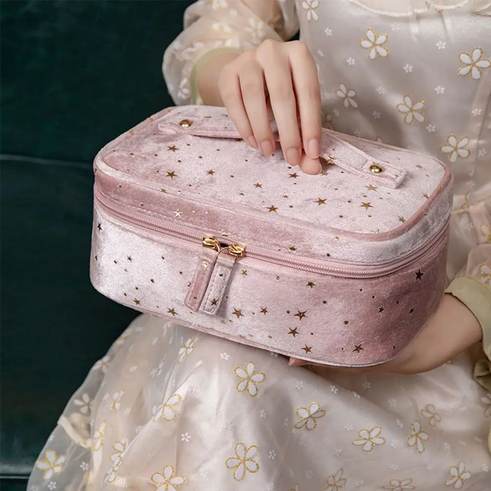 Astuccio cosmetico rosa in velluto con stelle dorate, chiusura a zip, tenuto da una mano femminile. Elegante e spazioso, ideale per viaggi.
