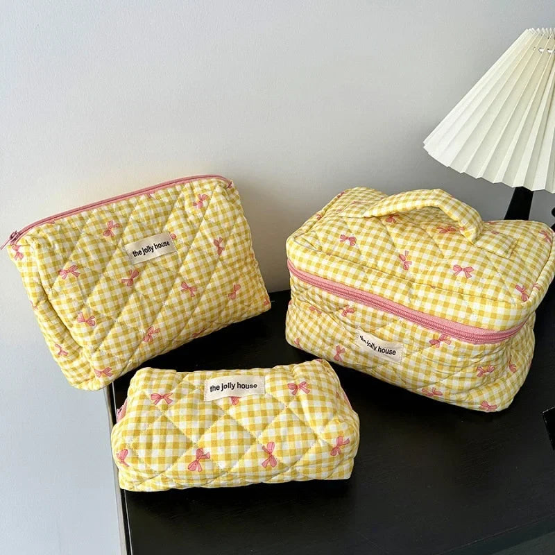 Set di tre beauty case gialli a quadretti con fiocchi rosa, su tavolo nero. Include borsa con manico, pochette e astuccio. Design elegante e pratico.