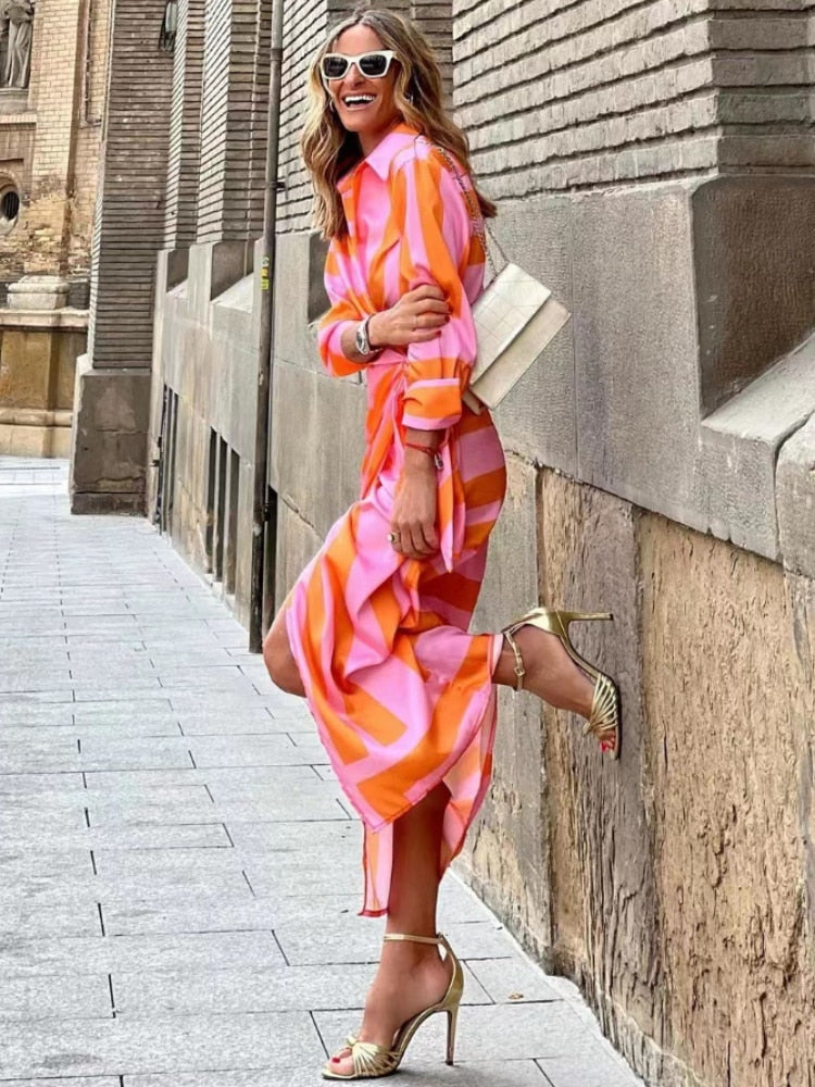 Donna sorridente in abito lungo colorato arancione e rosa, occhiali da sole bianchi, sandali dorati, posa su marciapiede urbano. Moda estiva elegante.