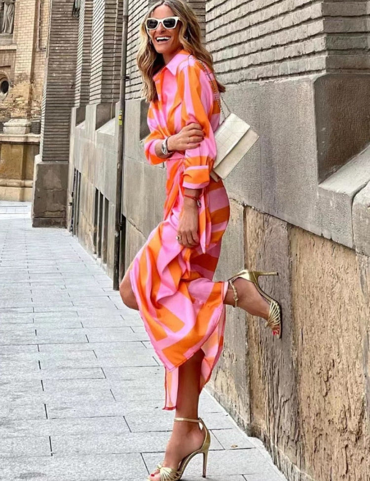 Donna sorridente in abito lungo colorato arancione e rosa, occhiali da sole bianchi, sandali dorati, posa su marciapiede urbano. Moda estiva elegante.