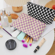 Set di trucchi su tavolo bianco con pochette a scacchi, pennelli colorati, fondotinta, e accessori cosmetici. Organizzazione trucco elegante e pratica.