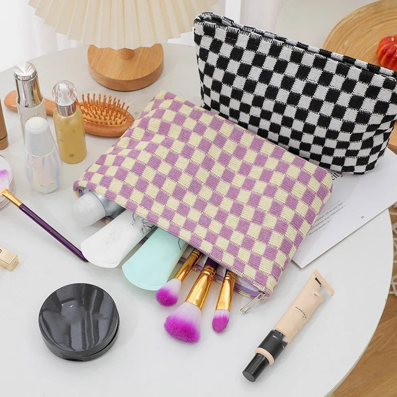 Set di trucchi su tavolo bianco con pochette a scacchi, pennelli colorati, fondotinta, e accessori cosmetici. Organizzazione trucco elegante e pratica.