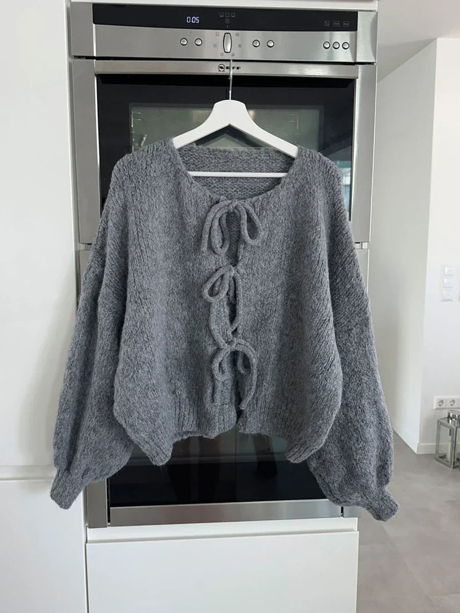 Maglione grigio in lana con lacci decorativi, appeso su gruccia davanti a forno moderno. Design casual, ideale per l'autunno e l'inverno.