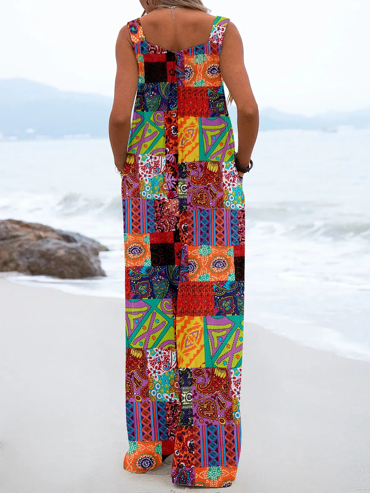 Donna in spiaggia con tuta lunga patchwork colorata, design geometrico e floreale, moda estiva, abbigliamento casual, mare sullo sfondo.