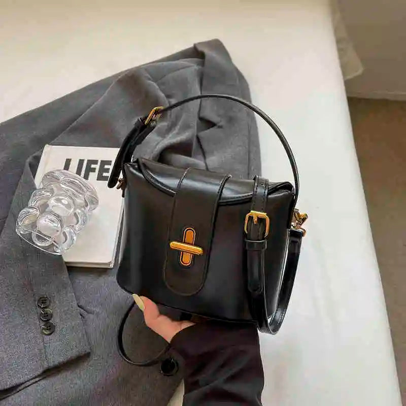 Borsa a tracolla in pelle nera con fibbia dorata, posata su giacca grigia e rivista. Elegante accessorio moda donna, stile classico e versatile.