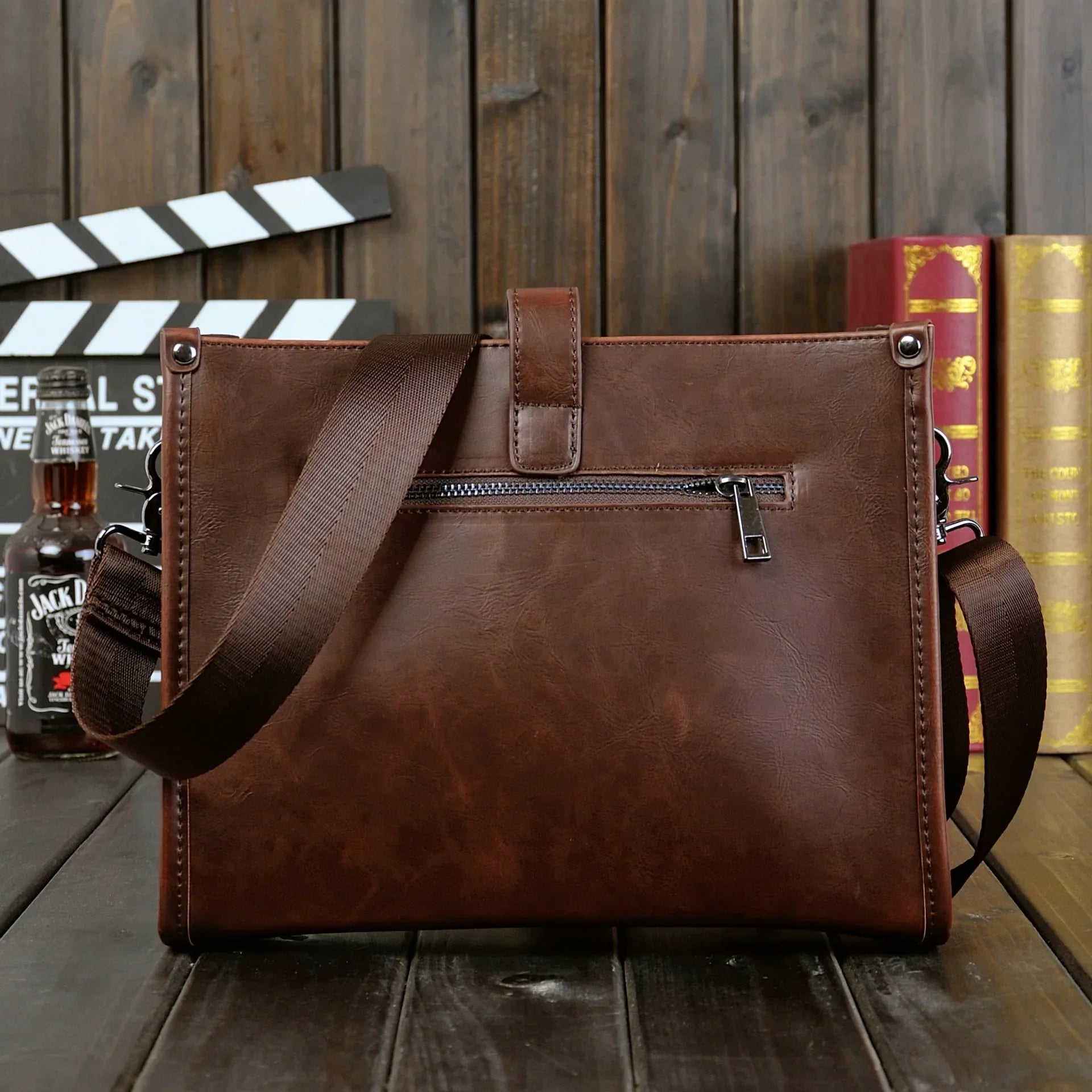Borsa a tracolla in pelle marrone con zip, design elegante, ideale per lavoro e viaggio. Sfondo con libri e bottiglia. Accessorio moda uomo.