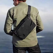 Uomo con borsa a tracolla nera all'aperto, giacca verde, vista mare. Accessorio moda maschile, borsa resistente, stile casual.
