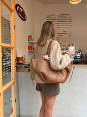 Donna in caffetteria con borsa a spalla marrone, cappotto beige e caffè in mano. Interno moderno con menu a parete. Shopping, moda, accessori.