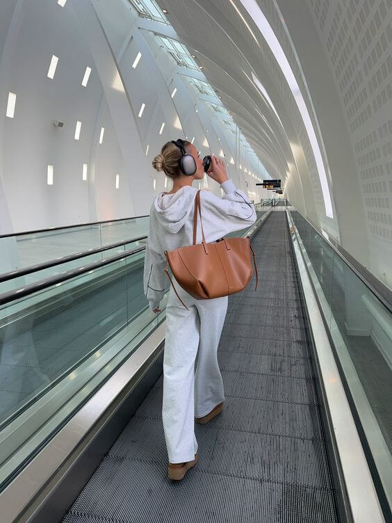 Donna in tuta grigia e cuffie cammina su scala mobile in aeroporto moderno, borsa in pelle marrone. Viaggio, moda, comfort.