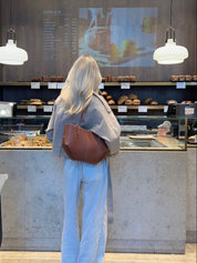 Donna bionda in caffetteria, indossa pantaloni a righe e giacca grigia, osserva il menu sopra il bancone con dolci e pane esposti.