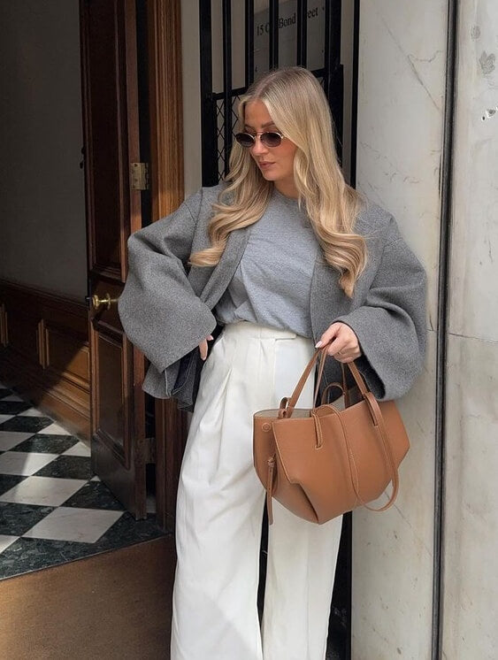 Donna con occhiali da sole indossa cappotto grigio oversize, maglia grigia e pantaloni bianchi, tenendo borsa marrone. Moda autunnale elegante.