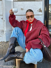 Donna con giacca rossa oversize, jeans larghi, stivali neri e occhiali da sole, tiene una tazza di caffè. Look moda street style autunno.