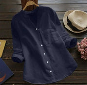 Camicia blu scuro da donna in lino, maniche lunghe, colletto alla coreana, su sfondo di legno con cappello di paglia e fiori rosa.