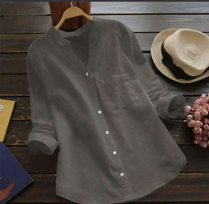 Camicia grigia in lino con colletto alla coreana, maniche lunghe arrotolate, su tavolo di legno scuro accanto a cappello di paglia e fiori rosa.