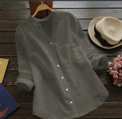 Camicia grigia in lino con colletto alla coreana, maniche lunghe arrotolate, su tavolo di legno scuro accanto a cappello di paglia e fiori rosa.