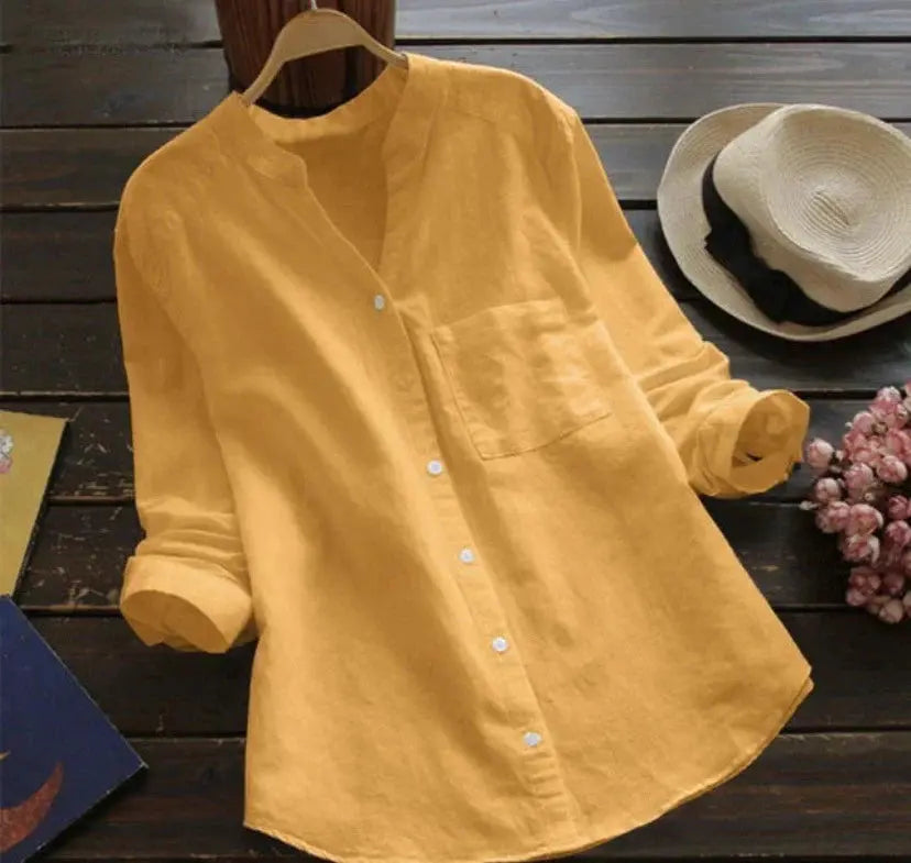 Camicia gialla in lino con colletto alla coreana, maniche lunghe arrotolate, appesa su gruccia. Sfondo con cappello di paglia e fiori rosa.