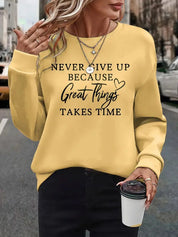Donna con felpa gialla con scritta motivazionale, collane a strati, pantaloni neri, tiene bicchiere di caffè. Moda casual autunnale urbana.