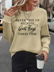 Donna con maglione beige con scritta motivazionale, collane a strati, pantaloni neri, tiene una tazza da caffè. Moda casual autunnale urbana.