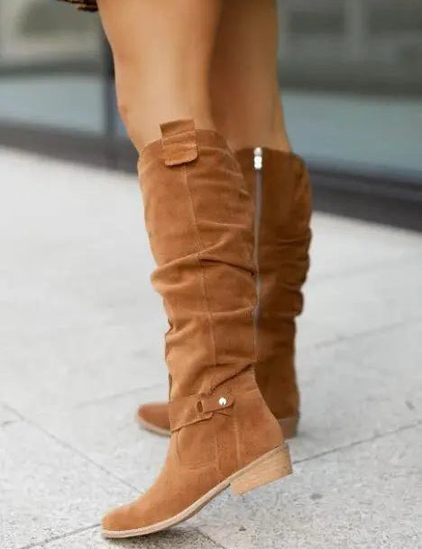 Stivali alti in camoscio marrone chiaro, tacco basso, indossati da una persona su marciapiede. Moda autunnale, calzature eleganti donna.
