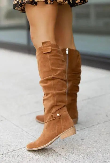 Stivali alti in camoscio marrone chiaro, tacco basso, indossati da una persona su marciapiede. Moda autunnale, calzature eleganti donna.